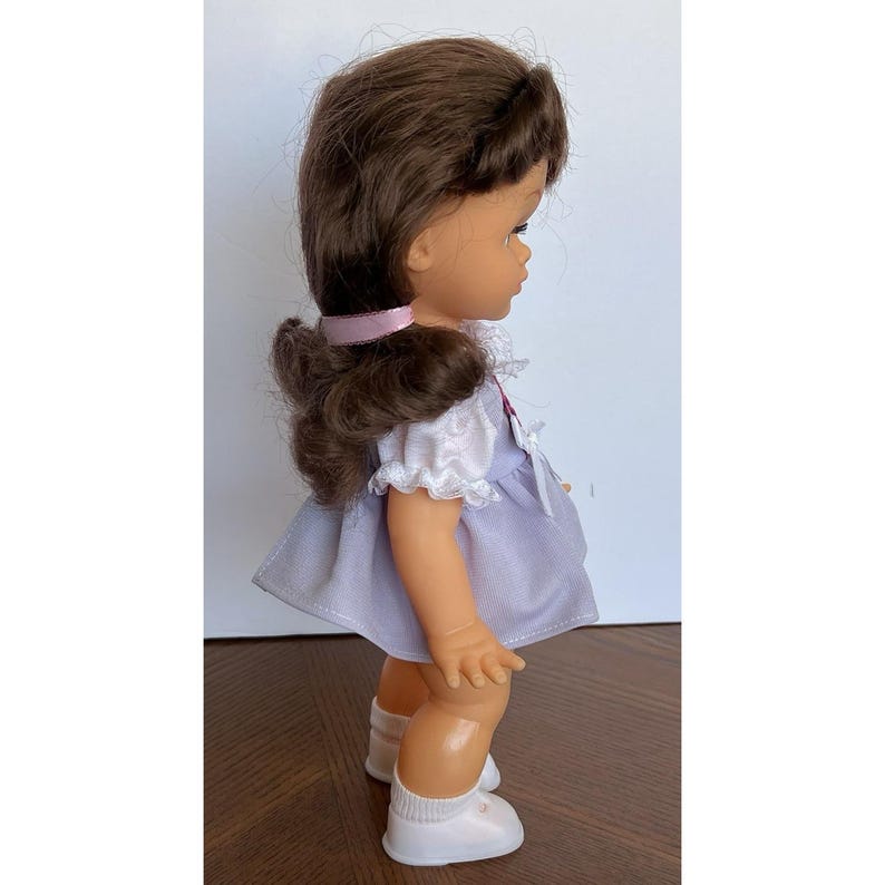 Peut inclure: Poup&eacute;e vintage aux cheveux bruns attach&eacute;s en queue de cheval, v&ecirc;tue d'une robe violet clair avec une bordure en dentelle blanche. La poup&eacute;e porte des chaussures blanches et un ruban rose. La poup&eacute;e est sur une surface en bois.