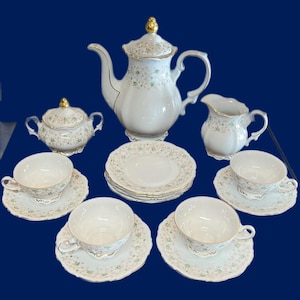 May include: A complete white porcelain tea set with gold trim and delicate floral patterns. The set includes a teapot, sugar bowl, creamer, saucers, and teacups. The teapot is approximately 10 inches tall.