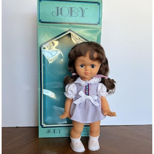 Peut inclure: Poup&eacute;e vintage nomm&eacute;e Joey, avec des cheveux bruns et des yeux bleus, v&ecirc;tue d'une robe lavande avec un col et un n&oelig;ud blancs. La poup&eacute;e est dans sa bo&icirc;te d'origine, sur laquelle est imprim&eacute; le nom "JOEY".