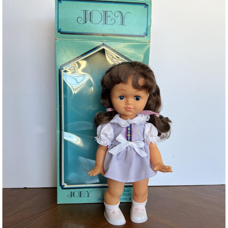 Peut inclure: Poup&eacute;e vintage nomm&eacute;e Joey, avec des cheveux bruns et des yeux bleus, v&ecirc;tue d'une robe lavande avec un col et un n&oelig;ud blancs. La poup&eacute;e est dans sa bo&icirc;te d'origine, sur laquelle est imprim&eacute; le nom "JOEY".