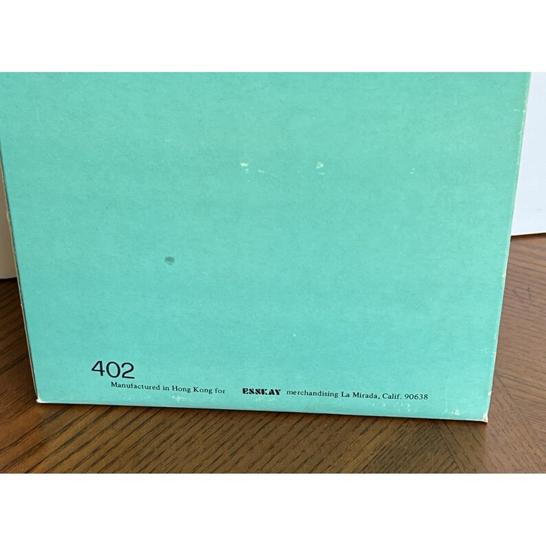 Peut inclure: Une section d'une bo&icirc;te avec une surface de couleur sarcelle. Le num&eacute;ro "402" est imprim&eacute; dans le coin inf&eacute;rieur gauche. En dessous, le texte indique "Manufactured in Hong Kong for ESSKAY merchandising La Mirada, Calif. 90638."