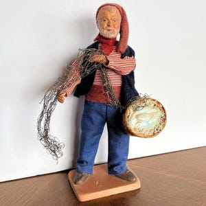 May include: A figurine of an elderly fisherman wearing a red and white striped shirt, blue pants, and a long red cap. He holds a fishing net and a basket with a fish inside. The figurine stands on a terracotta base.
