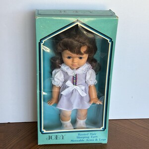 Peut inclure: Poup&eacute;e vintage Joey dans une bo&icirc;te turquoise. La poup&eacute;e a des cheveux bruns et des yeux bleus, v&ecirc;tue d'une robe lavande avec un col et un n&oelig;ud blancs. La bo&icirc;te indique "Rooted Hair, Sleeping Eyes, Moveable Arms & Legs."