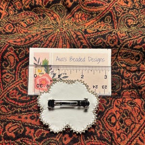 May include: A white, cloud-shaped hair clip with a silver metal clasp on the back, featuring a beaded edge. A ruler placed behind the clip shows it measures approximately 2 inches in width. A card labeled Ava's Beaded Designs is visible.