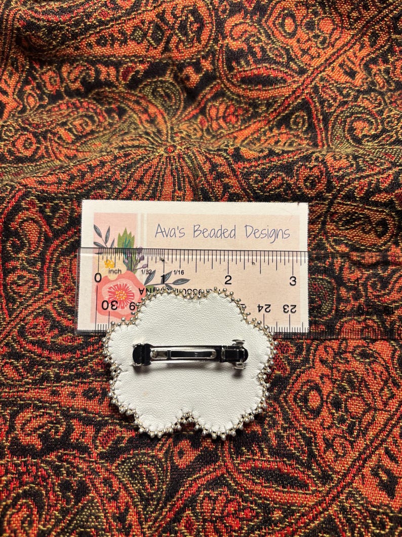 May include: A white, cloud-shaped hair clip with a silver metal clasp on the back, featuring a beaded edge. A ruler placed behind the clip shows it measures approximately 2 inches in width. A card labeled Ava's Beaded Designs is visible.