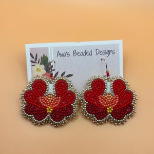 May include: A pair of flower-shaped beaded earrings featuring red, dark red, and peach beads with a yellow center, outlined in metallic silver beads. The earrings are attached to a white display card printed with the text Ava's Beaded Designs.