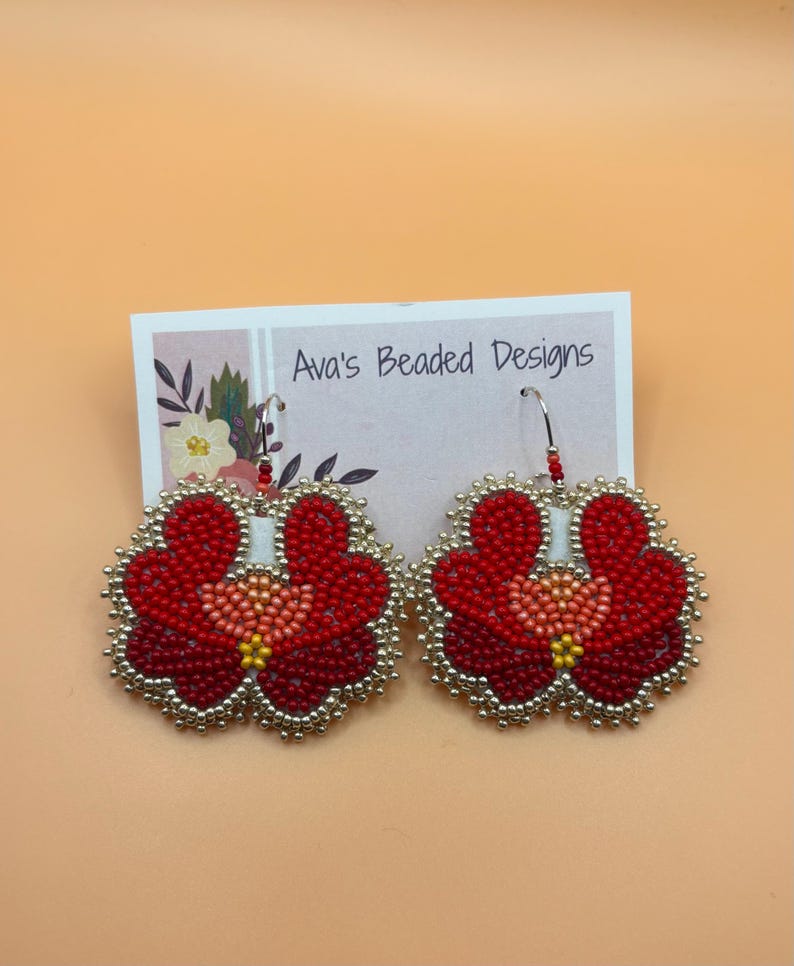 May include: A pair of flower-shaped beaded earrings featuring red, dark red, and peach beads with a yellow center, outlined in metallic silver beads. The earrings are attached to a white display card printed with the text Ava's Beaded Designs.