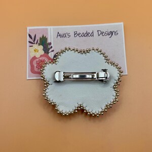 May include: A flower-shaped brooch with a white leather backing and a silver-toned metal bar pin mechanism. The edge is decorated with small gold-colored beads. A rectangular display card behind the brooch features the text Ava's Beaded Designs.