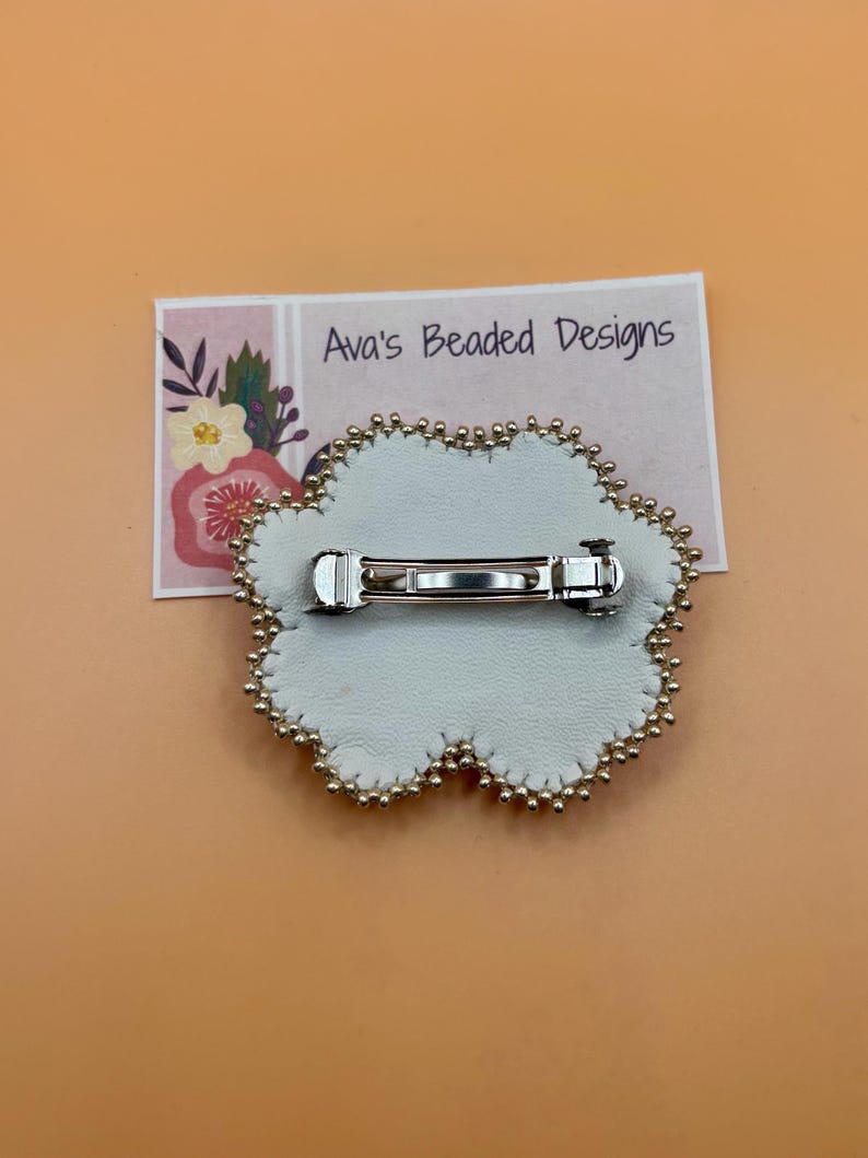 May include: A flower-shaped brooch with a white leather backing and a silver-toned metal bar pin mechanism. The edge is decorated with small gold-colored beads. A rectangular display card behind the brooch features the text Ava's Beaded Designs.