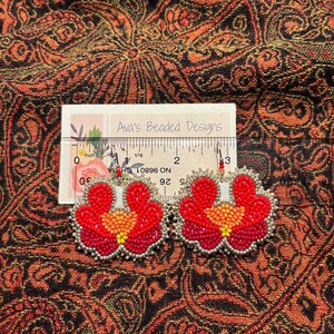 May include: A pair of floral-shaped beaded earrings featuring red, orange, and yellow seed beads with silver-colored edging. A ruler placed behind the earrings shows they measure approximately 2.5 inches in width. The brand name Ava's Beaded Designs is printed on the white card.