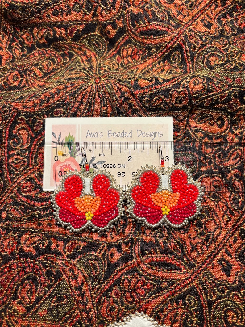 May include: A pair of floral-shaped beaded earrings featuring red, orange, and yellow seed beads with silver-colored edging. A ruler placed behind the earrings shows they measure approximately 2.5 inches in width. The brand name Ava's Beaded Designs is printed on the white card.