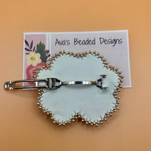 May include: A white flower-shaped hair clip with a silver-toned metal barrette mechanism on the back. The edges of the white material are lined with small gold-colored beads. A rectangular card behind the clip displays the text Ava's Beaded Designs.