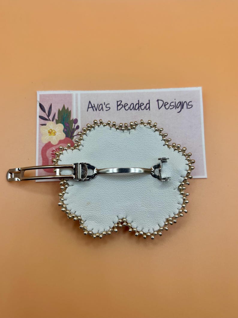 May include: A white flower-shaped hair clip with a silver-toned metal barrette mechanism on the back. The edges of the white material are lined with small gold-colored beads. A rectangular card behind the clip displays the text Ava's Beaded Designs.