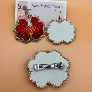 May include: A pair of flower-shaped beaded earrings and a matching hair clip on a display card labeled Ava's Beaded Designs. The earrings feature red, orange, and yellow beads with silver edging. The hair clip has a white leather backing and a silver metal barrette.