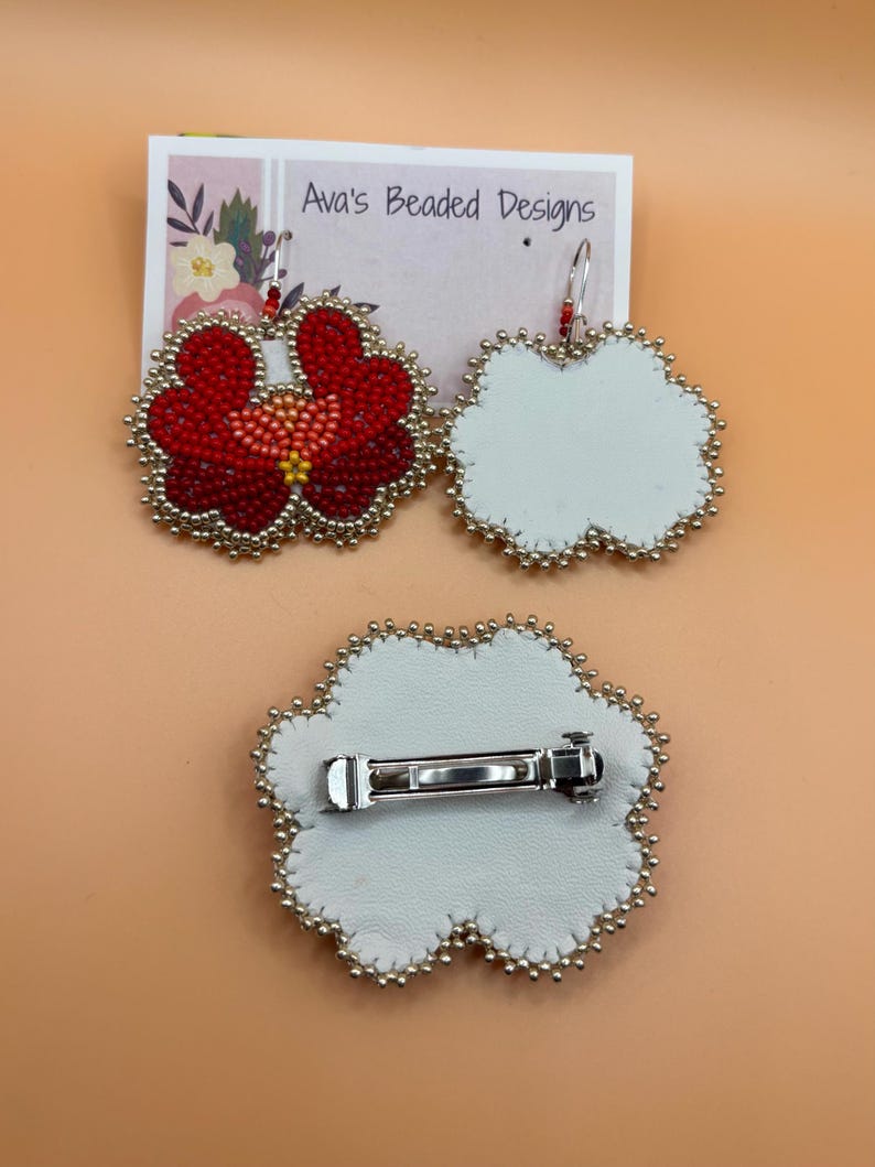 May include: A pair of flower-shaped beaded earrings and a matching hair clip on a display card labeled Ava's Beaded Designs. The earrings feature red, orange, and yellow beads with silver edging. The hair clip has a white leather backing and a silver metal barrette.