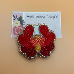 May include: A handcrafted beaded flower brooch featuring vibrant red petals, a peach-colored center, and yellow accents, all outlined with metallic silver beads. The brooch is displayed on a white card printed with the text Ava's Beaded Designs.