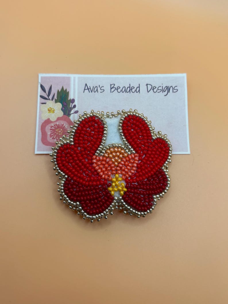May include: A handcrafted beaded flower brooch featuring vibrant red petals, a peach-colored center, and yellow accents, all outlined with metallic silver beads. The brooch is displayed on a white card printed with the text Ava's Beaded Designs.