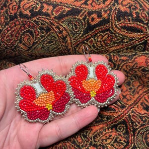 May include: A pair of dangle earrings featuring a floral design crafted from small beads in shades of bright red, deep burgundy, orange, and yellow. The edges are outlined with metallic silver beads, and each earring hangs from a silver hook.