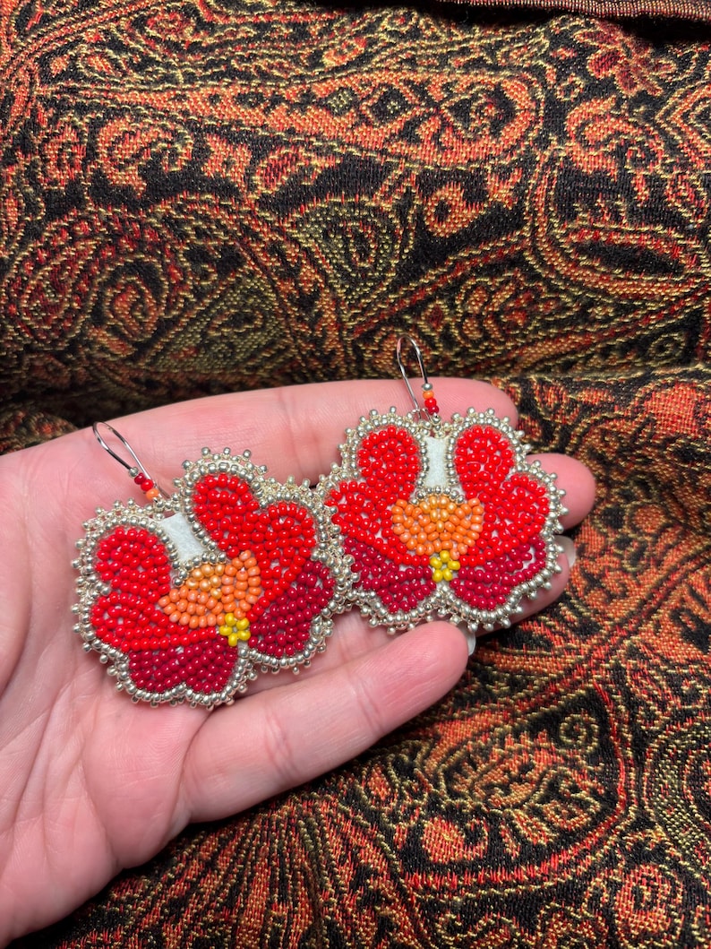 May include: A pair of dangle earrings featuring a floral design crafted from small beads in shades of bright red, deep burgundy, orange, and yellow. The edges are outlined with metallic silver beads, and each earring hangs from a silver hook.
