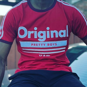 May include: Red t-shirt with white stripes on the sleeves and a white graphic that says "Original Pretty Boys" with a white stripe underneath.