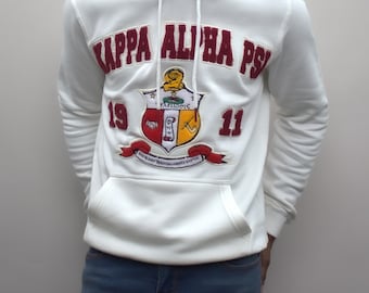 Może przedstawiać: Biała bluza z kapturem z haftowanym herbem w kolorze bordowym i złotym, na którym widnieje napis "Kappa Alpha Psi" i cyfry "1911".