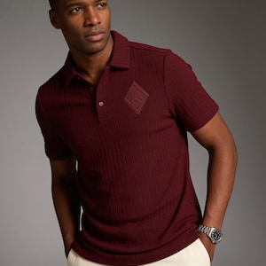 May include: A short-sleeved, ribbed polo shirt in a deep burgundy color. The shirt features a classic collar, a buttoned placket, and a tonal diamond-shaped patch on the left chest. The fabric has a vertical textured pattern throughout the garment.