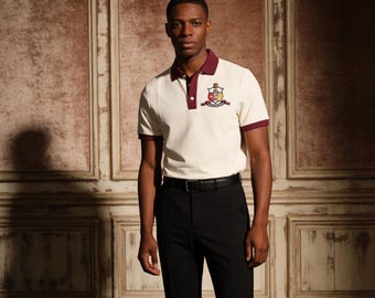 Op de afbeelding: Crèmekleurige poloshirt met bordeauxrode kraag en manchetten, met een embleem op de borst. Gedragen met een zwarte broek en een zwarte riem. De achtergrond is een getextureerde, lichtbruine muur.