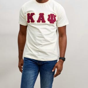 May include: Cream-colored t-shirt with maroon Greek letters "KAΨ" across the chest. The shirt is paired with blue jeans. The t-shirt has short sleeves and a crew neck.