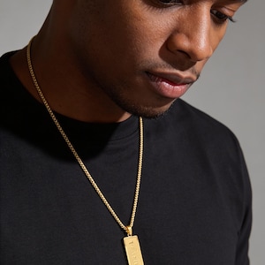 May include: A gold-toned metal necklace featuring a thin curb chain and a rectangular pendant. The pendant has a vertical orientation and is engraved with the text "STRENGTH" in a clear, sans-serif font, centered on the polished metal surface.