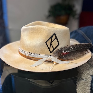 May include: A cream colored cowboy hat with a black diamond logo and a brown leather band. A brown feather is attached to the hat.