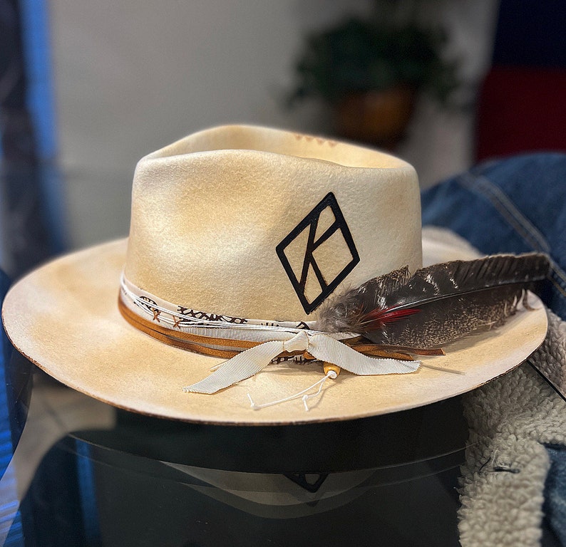 May include: A cream colored cowboy hat with a black diamond logo and a brown leather band. A brown feather is attached to the hat.