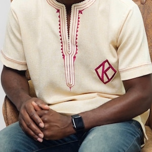 Pode incluir: Camisa creme de manga curta com bordados bordô ao redor da gola e um emblema em forma de diamante. A camisa tem um design de decote em V e é feita de um tecido texturizado. A pessoa está usando jeans azuis e um relógio preto.