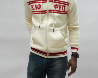 May include: Cream-colored zip-up hoodie with red stripes and Greek letters. The hoodie has a hood, front pockets, and red trim on the cuffs and waist. The person is wearing blue jeans.