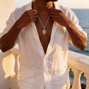 May include: A white short-sleeved button-down shirt with a textured fabric finish and a chest pocket. A silver-toned chain necklace features a diamond-shaped pendant with a cutout letter K design, resting against the chest area of the shirt.
