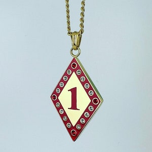 May include: A gold pendant necklace with a red diamond-shaped charm. The charm has a white background with the number "1" in red.