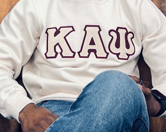 Puede incluir: Sudadera color crema con las letras griegas K, A y Ψ en burdeos y crema. Las letras están bordadas en la parte delantera de la sudadera. La sudadera es de manga larga y tiene cuello redondo.