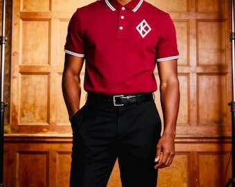Op de afbeelding: Een bordeauxrode poloshirt met witte details op de kraag en mouwen. Het shirt heeft een wit ruitvormig logo op de borst. Gedragen met een zwarte broek en een zwarte riem. De achtergrond is een houten wand.