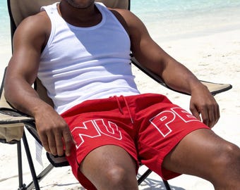 May include: A bright red bucket hat, a plain white ribbed vest, and red swim shorts featuring the white text "NU PE" printed across the front. The clothing is worn while seated in a folding outdoor chair with a tan fabric seat and black frame.