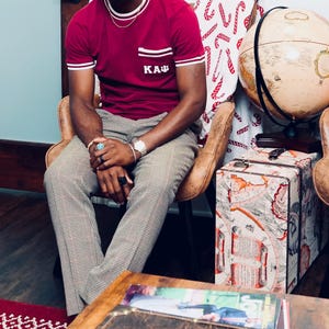 Könnte beinhalten: Burgunderfarbenes T-Shirt mit weißen Zierleisten und einer Tasche mit den Buchstaben "KAΨ". Das Shirt wird mit einer gemusterten Hose kombiniert. Ein Globus und ein dekorativer Koffer mit Kartenmuster sind im Hintergrund zu sehen.