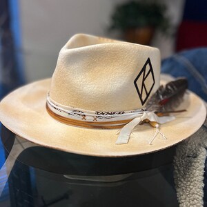 May include: A cream-colored felt fedora hat with a black diamond-shaped logo and a brown leather band with a white ribbon tied in a bow. A single feather is attached to the hat band.