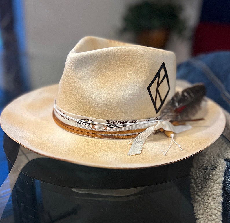 May include: A cream-colored felt fedora hat with a black diamond-shaped logo and a brown leather band with a white ribbon tied in a bow. A single feather is attached to the hat band.