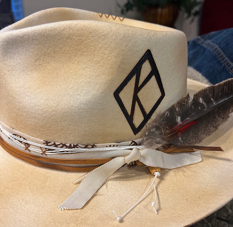 May include: A cream-colored felt hat with a black diamond-shaped logo and a brown leather band. A white ribbon is tied around the hat with a brown feather attached.