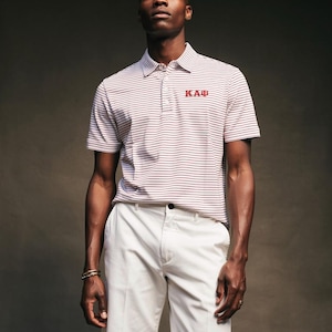 May include: A man wearing a striped polo shirt with the letters "KAB" in red and white shorts. The polo shirt has horizontal stripes in white and burgundy. The shorts are a solid white color.