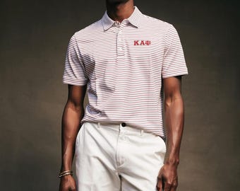 Op de afbeelding: Een man draagt een gestreepte poloshirt met de letters "KAB" in rood en een witte korte broek. De poloshirt heeft horizontale strepen in wit en bordeauxrood. De korte broek is effen wit.