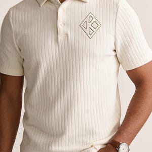 May include: A cream-colored short-sleeved polo shirt featuring a vertical ribbed texture, a classic collar, and a two-button placket. On the left chest, there is a dark embroidered geometric logo shaped like a diamond containing smaller shapes.