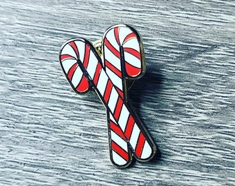 May include: A red and white candy cane enamel pin. The pin is shaped like two candy canes that are crossed over each other.