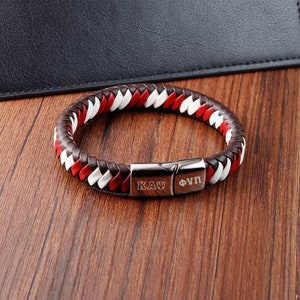 May include: A brown, white, and red braided leather bracelet with a silver clasp. The clasp has the text "ΚΛΥ ΦΥΠ" engraved on it.