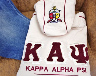 Może przedstawiać: Biała bluza z kapturem z herbem bractwa Kappa Alpha Psi w kolorze bordowym i białym oraz napisem "Kappa Alpha Psi Fraternity, Inc." w bordowych literach.
