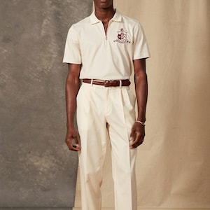 May include: A cream-colored polo shirt with a zipper and a crest emblem, paired with matching pleated trousers and a brown leather belt. The outfit is a classic, preppy style.