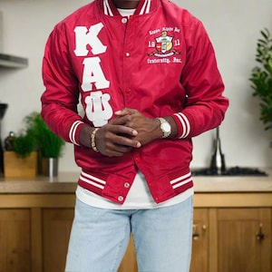 May include: Red satin bomber jacket with white stripes on the sleeves and collar. The jacket features the letters "KAU" in white on the front and a crest with text on the right chest. A classic varsity style.